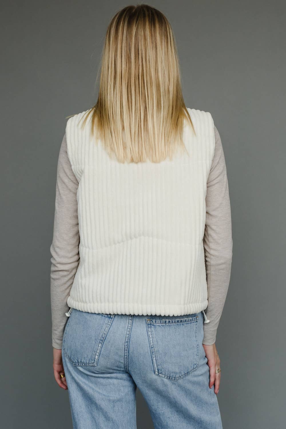Person wearing a beige ribbed vest over a long-sleeve shirt and blue jeans on a gray background