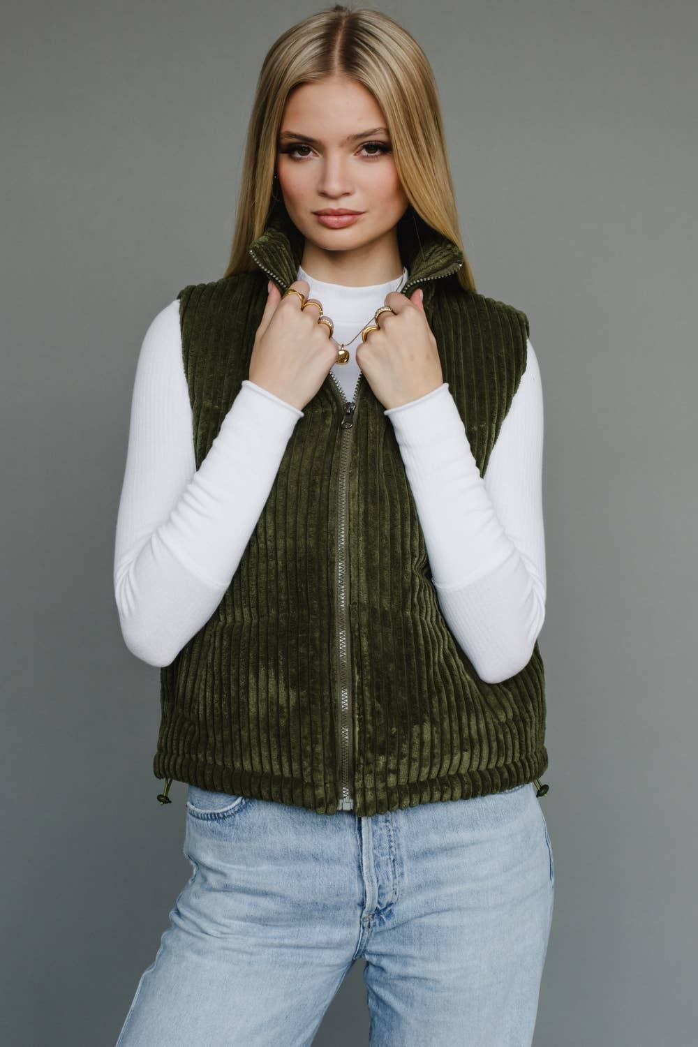 Woman wearing a green vest over a white long-sleeve shirt and blue jeans on a gray background
