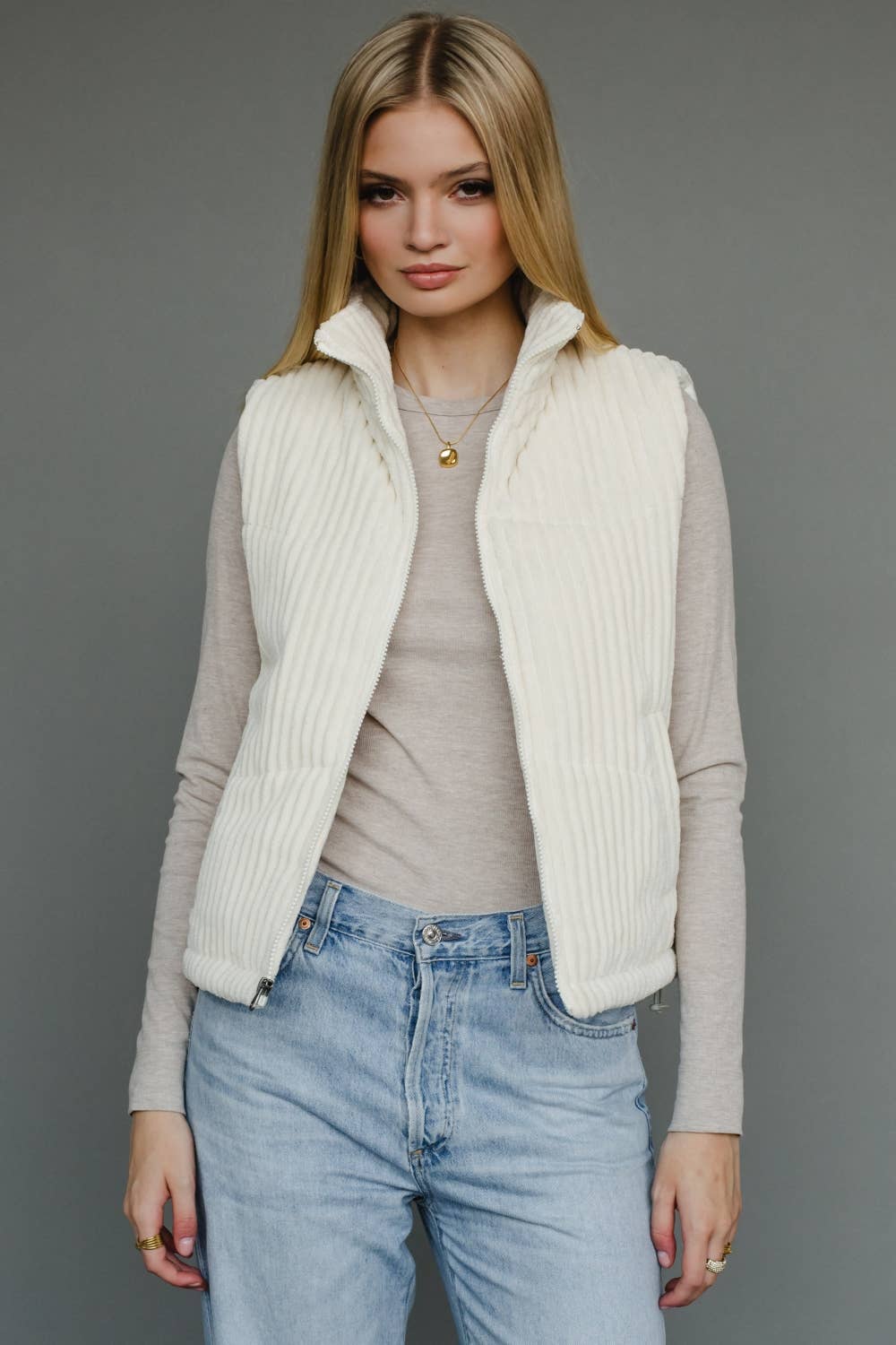 Woman wearing a cream vest over a gray sweater and blue jeans on a gray background
