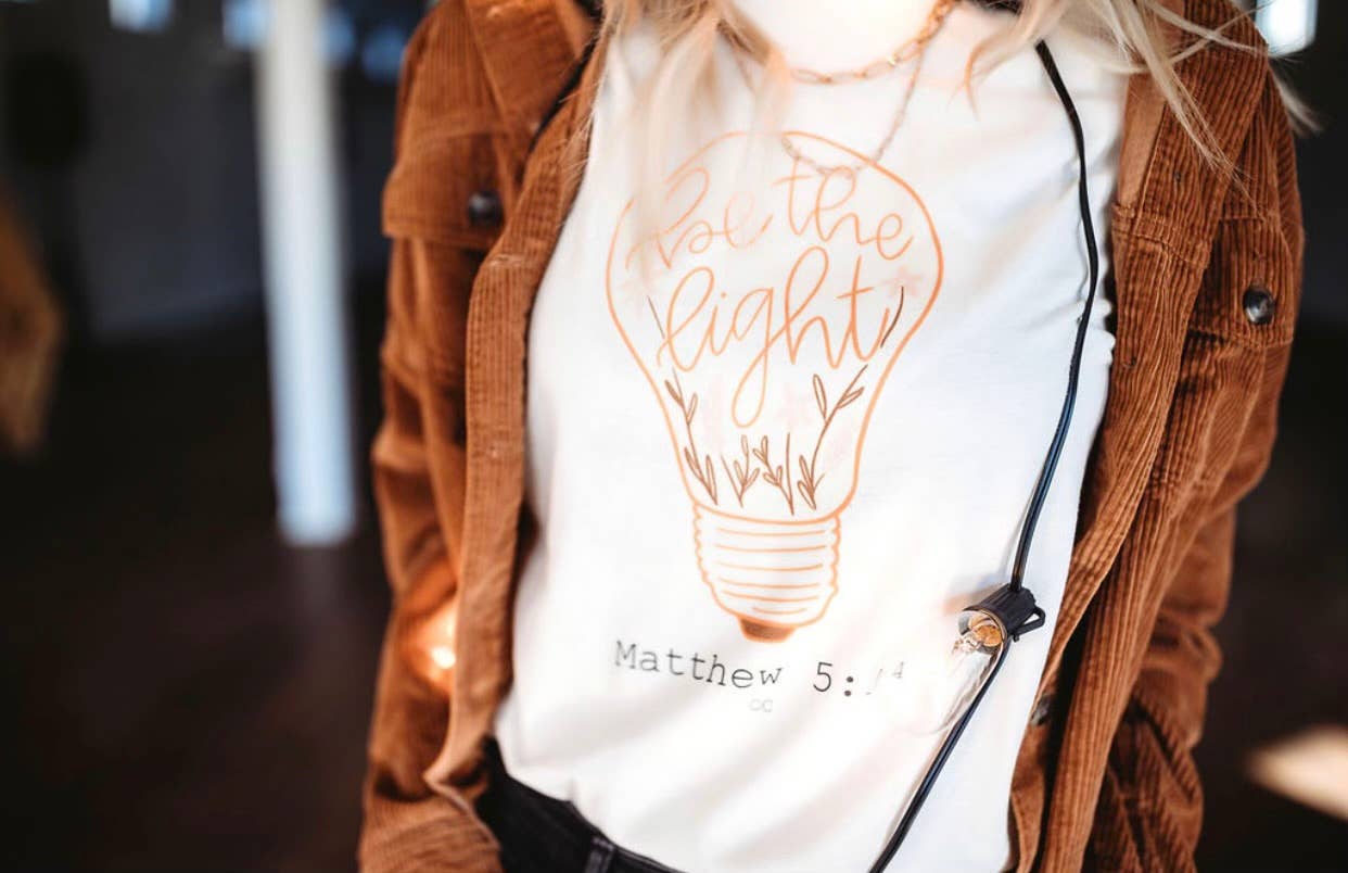 Person wearing a white t-shirt with a light bulb graphic and text, paired with a brown jacket.