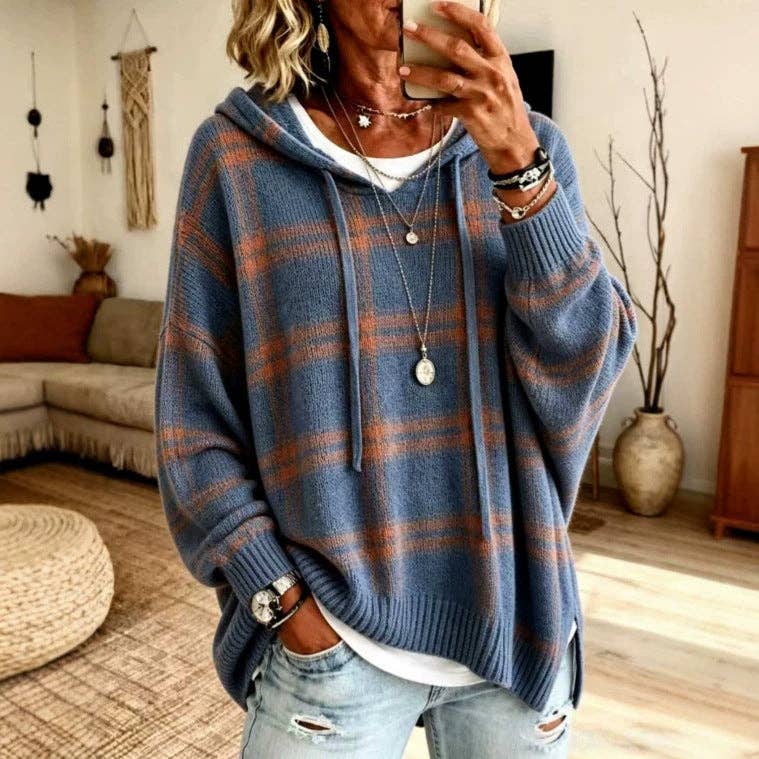 A woman takes a mirror selfie wearing an oversized blue and rust-orange plaid knit hoodie with a white undershirt and light-wash distressed jeans in a cozy living room setting.