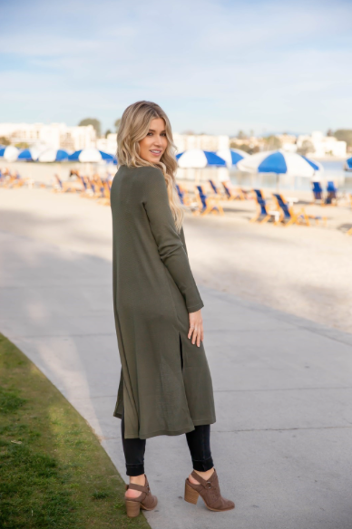 Woman in a long olive green dress standing on a sidewalk with beach umbrellas in the background.