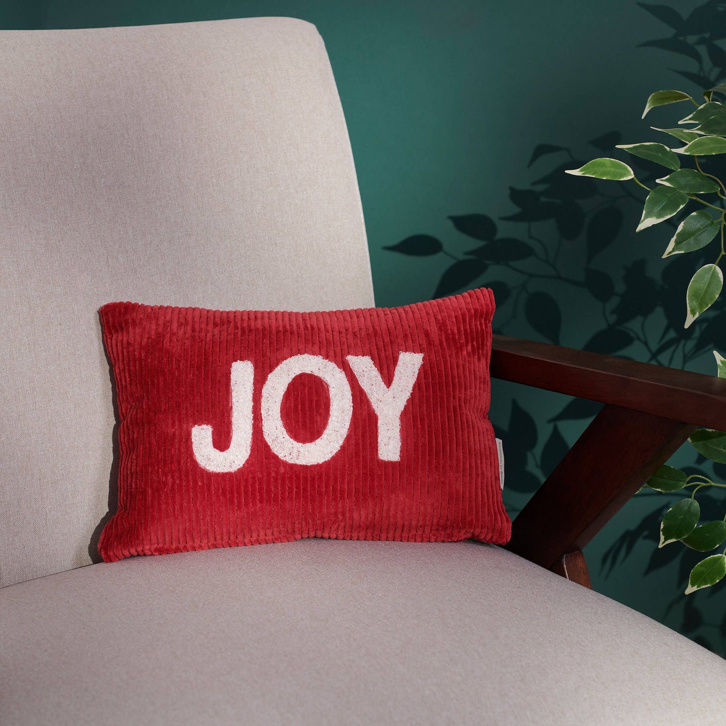 Red pillow with 'JOY' text on a gray chair against a teal wall with a plant.