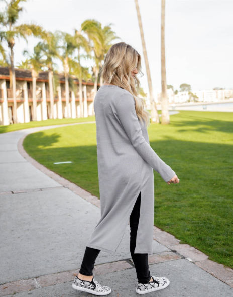 Woman wearing a long gray cardigan walking on a sidewalk with palm trees in the background