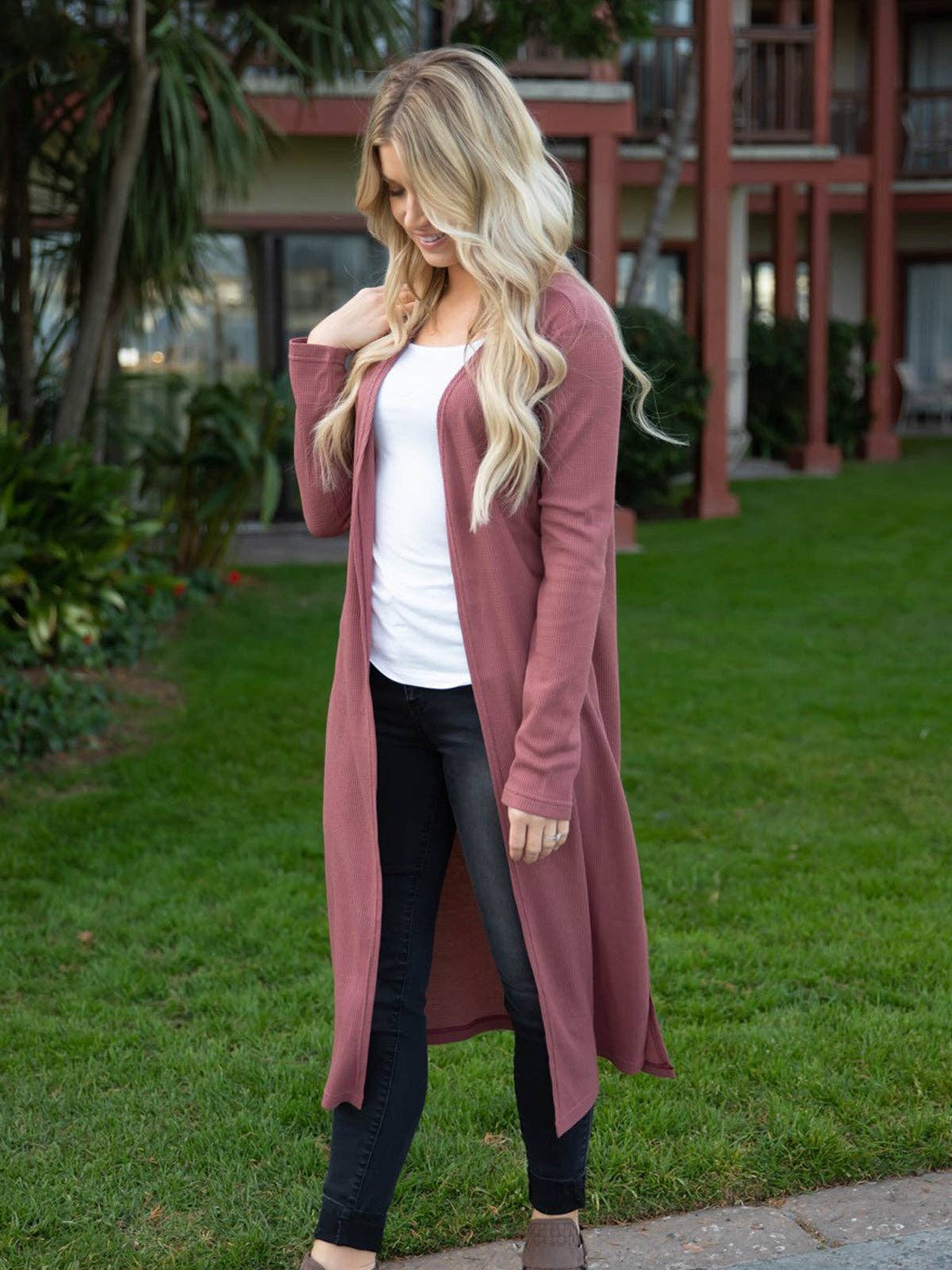 Woman wearing a long pink cardigan over a white top and black pants, standing outdoors.