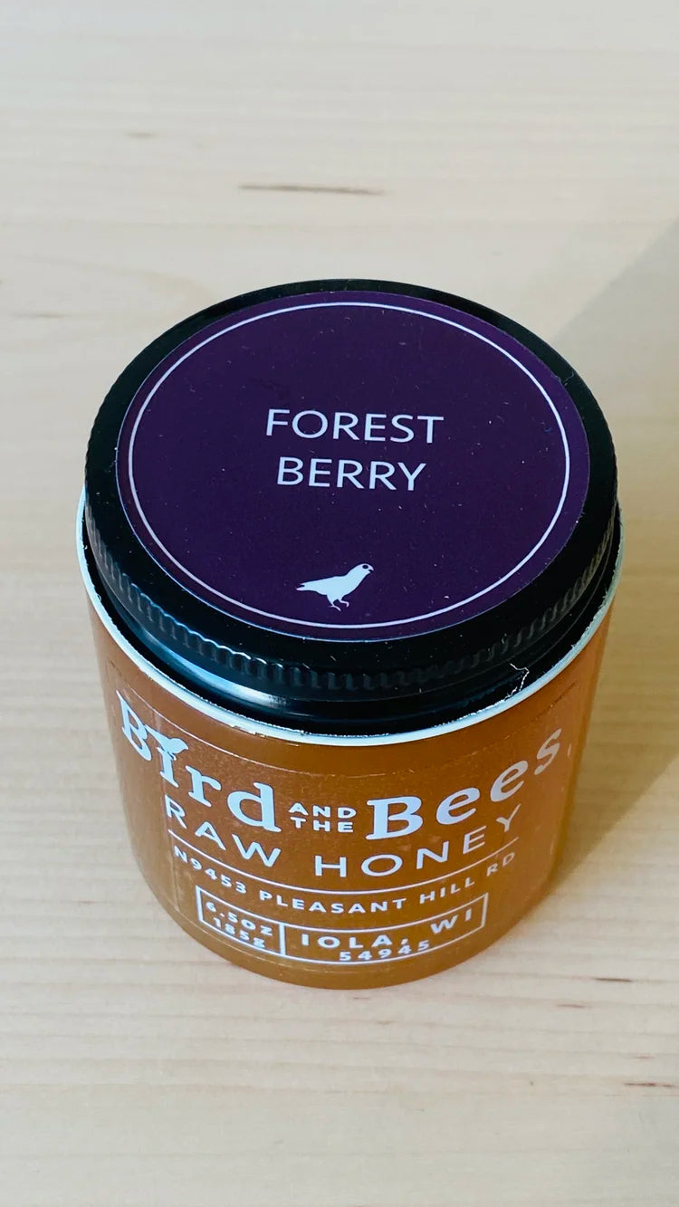 Jar of raw honey labeled 'Forest Berry' by 'Bird & The Bees' on a light wooden surface.