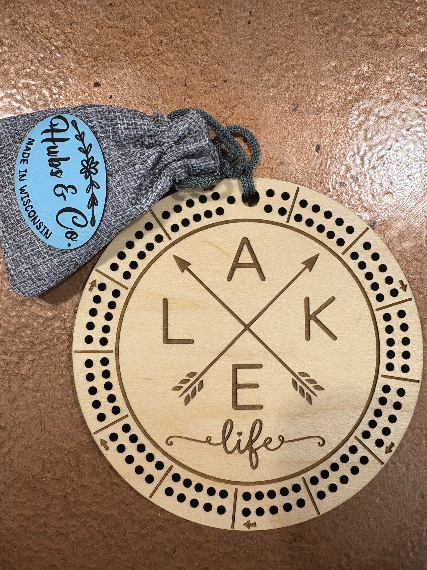 Wooden cribbage board with 'Lake Life' engraving and a gray drawstring bag with 'Hubs & Co.' label on a brown surface.