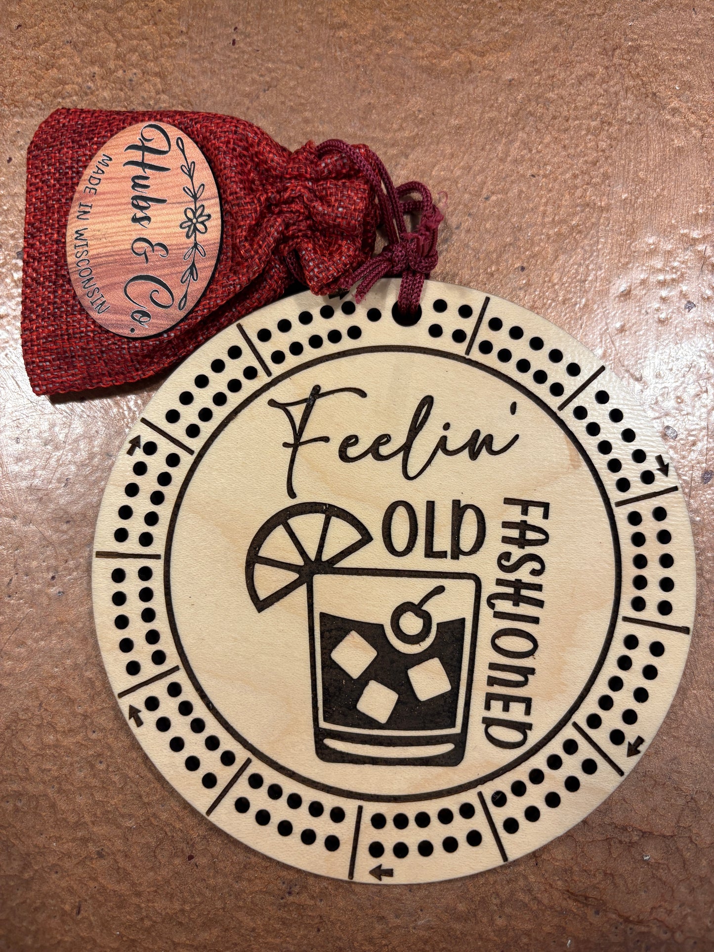 Round wooden cribbage with 'Fashioned Old Feeling' design and a small red bag with 'Hubs & Co.' branding on a brown surface.