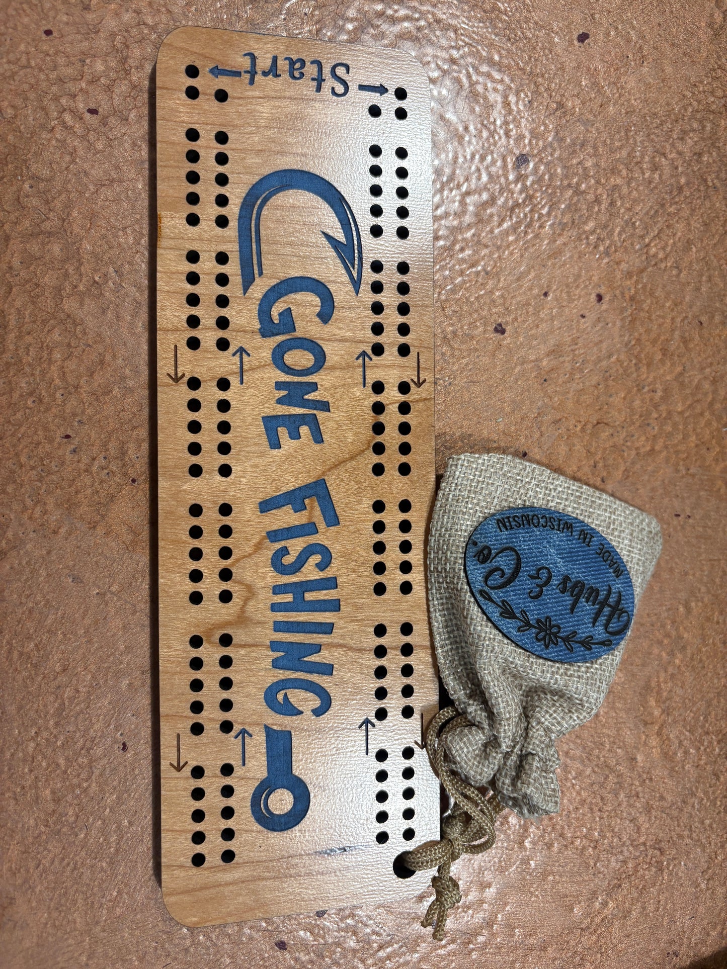 Wooden cribbage board with 'Gone Fishing' text and a drawstring bag on a textured surface