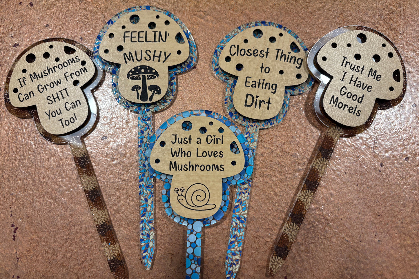 Five decorative mushroom-themed garden stakes with humorous sayings on a textured surface.