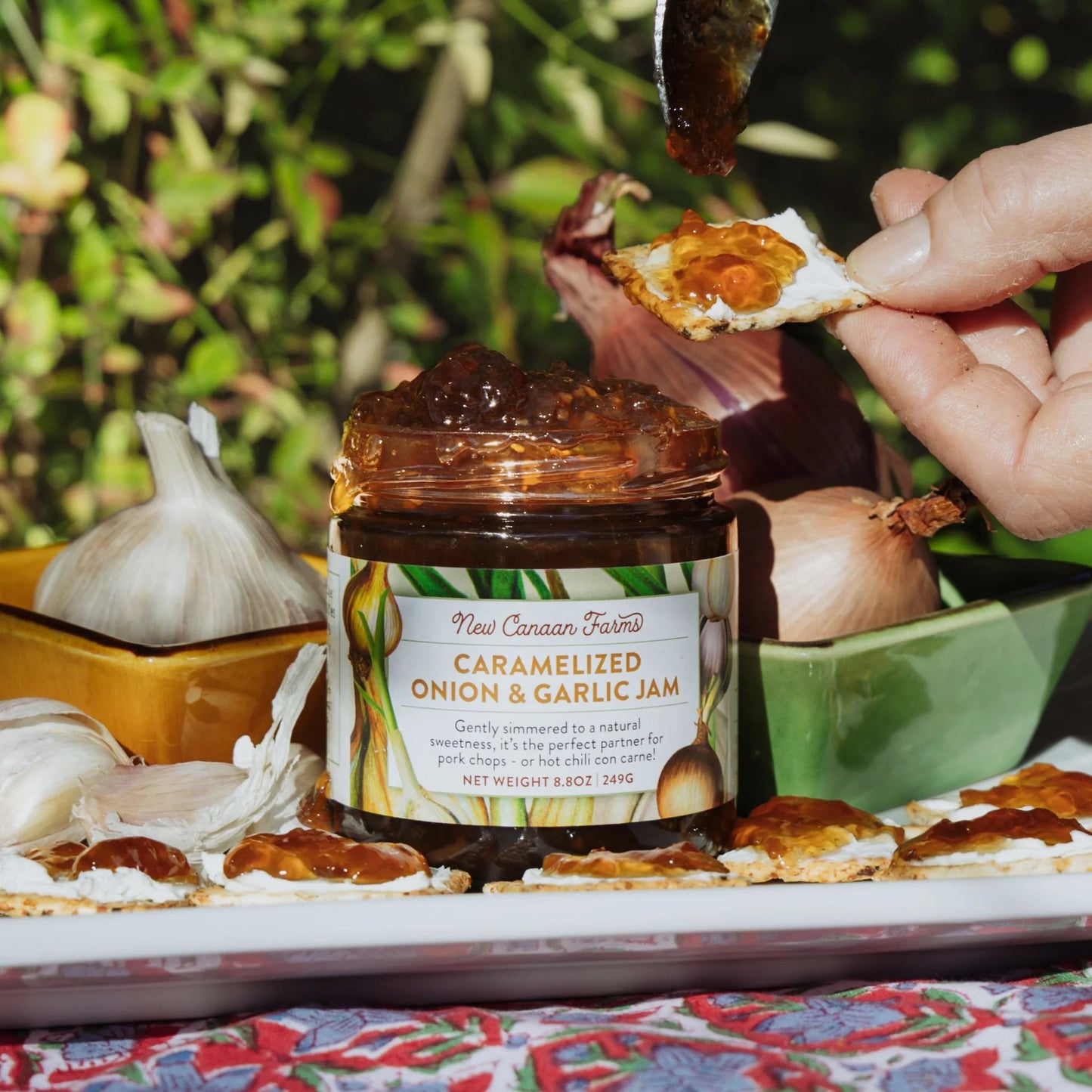 Person spreading caramelized onion and garlic jam on a cracker with a jar of the product in the foreground.