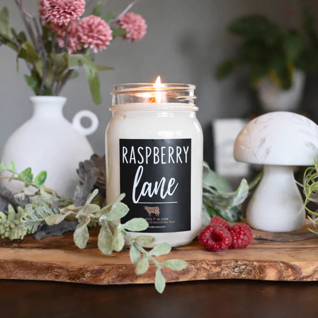Candle labeled 'Raspberry Lane' on a wooden surface with decorative elements.