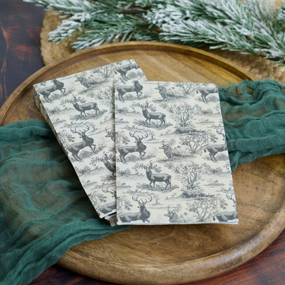 Three rectangular paper napkins with a classic toile de Jouy pattern of deer and trees on a cream background, styled on a round wooden tray with a green textured cloth and pine branches.