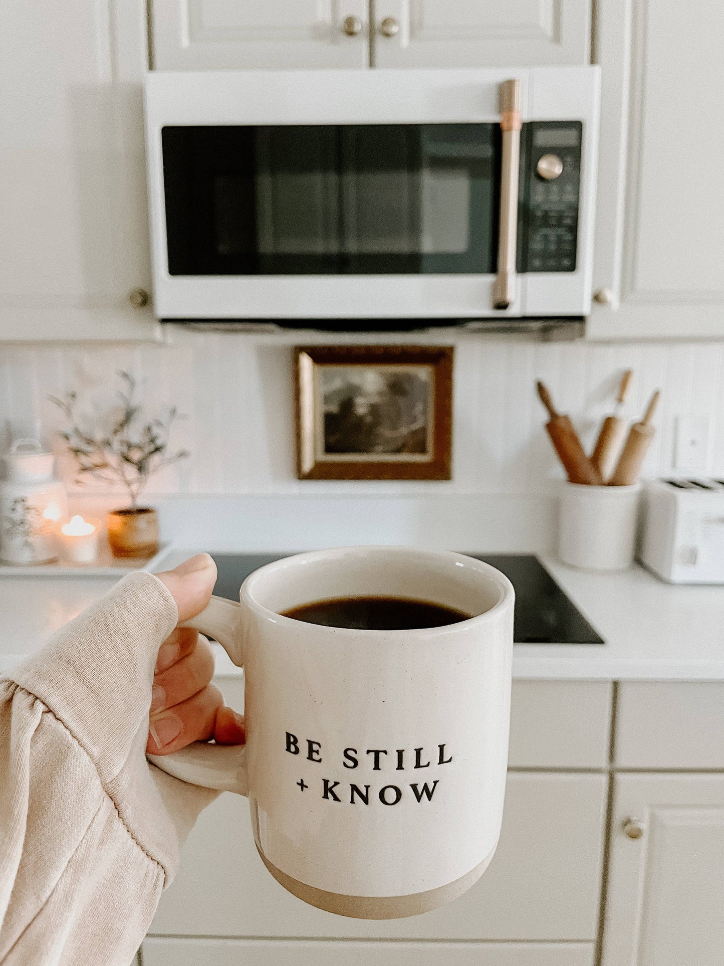 Person holding a mug with 'Be Still + Know' text in a kitchen.