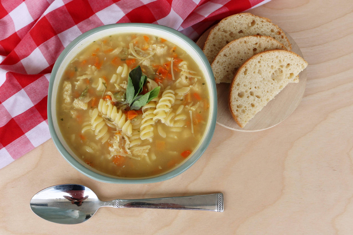 Bowl of soup with pasta and vegetables on a wooden table with bread slices and a checkered cloth.