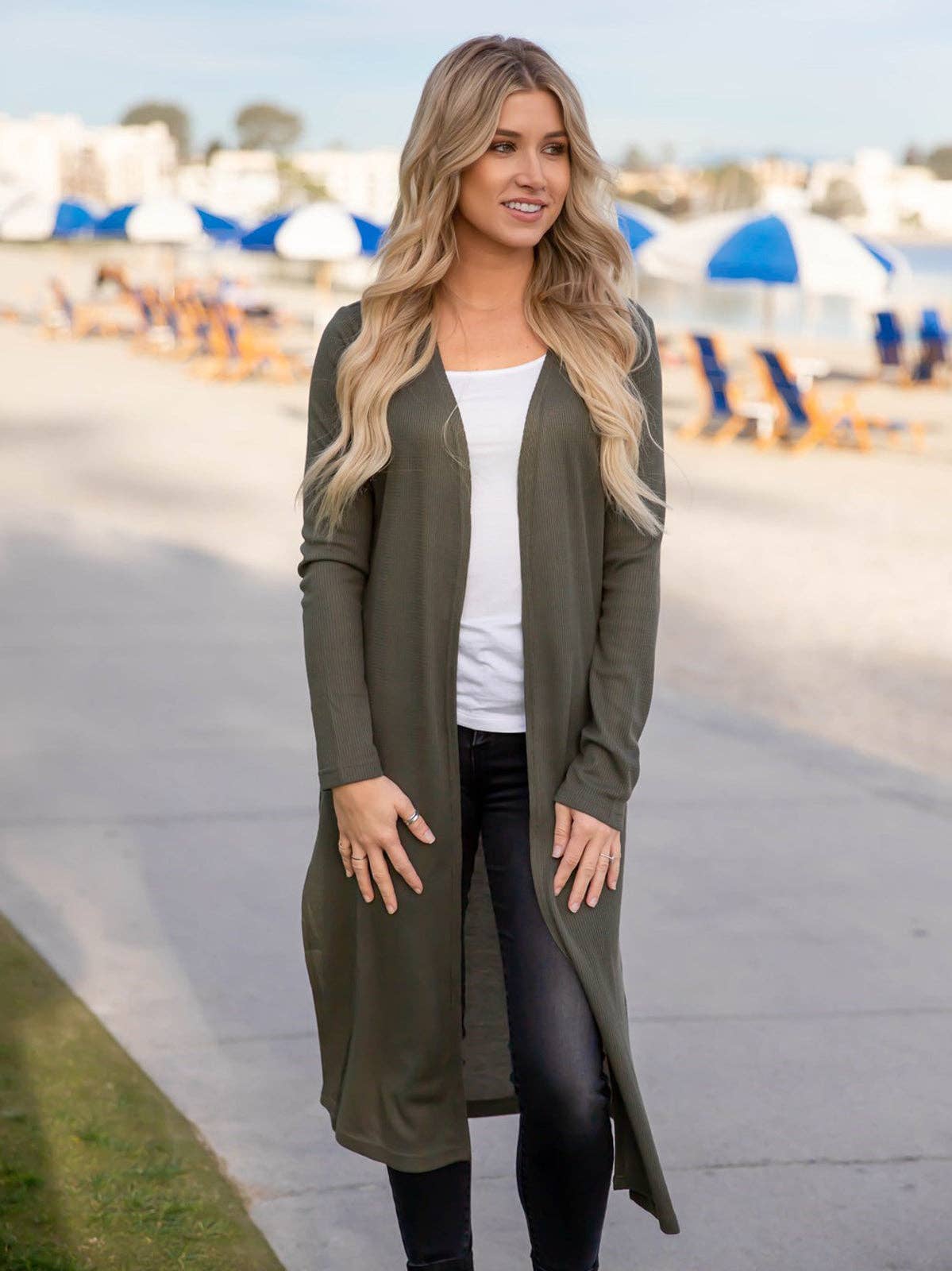 Woman wearing a long olive green cardigan over a white top and black pants, standing on a sidewalk with beach umbrellas in the background.