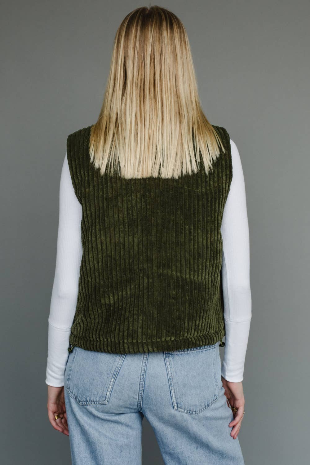 Person wearing a green knitted vest over a white long-sleeve shirt and blue jeans on a gray background