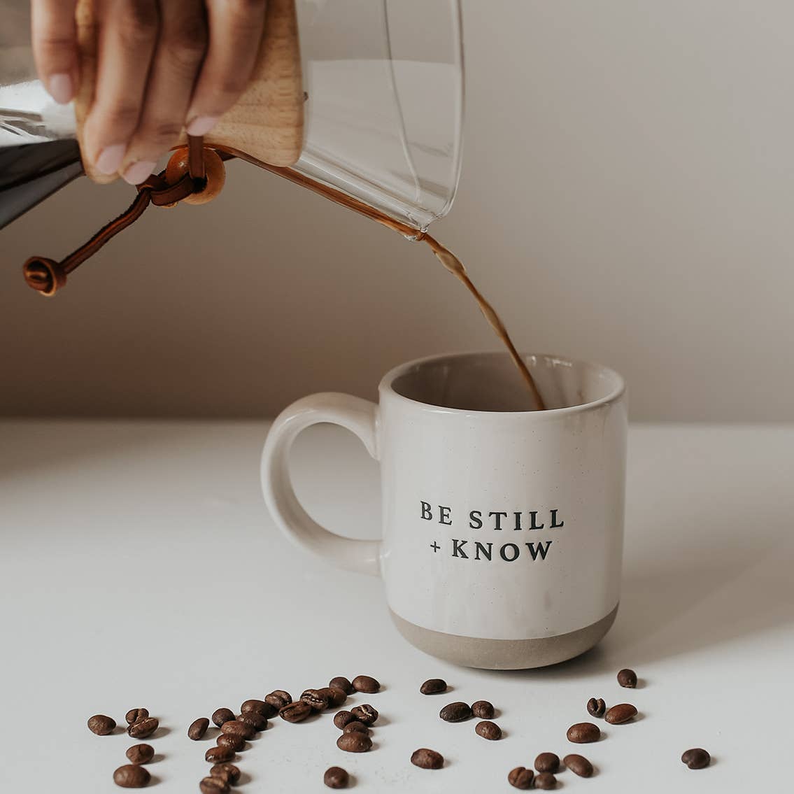 Person pouring coffee from a French press into a mug with 'Be Still + Know' text, surrounded by coffee beans.