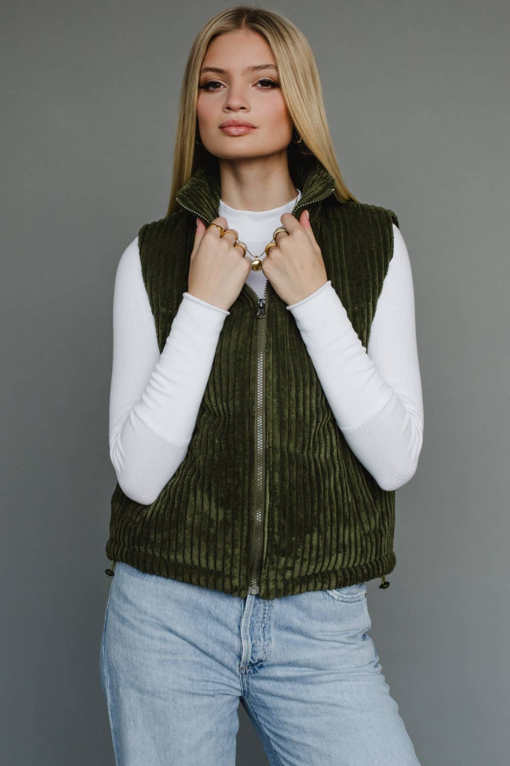Woman wearing a green vest over a white shirt and blue jeans on a gray background
