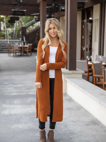 Woman wearing a long brown cardigan over a white top and black pants, standing outdoors.