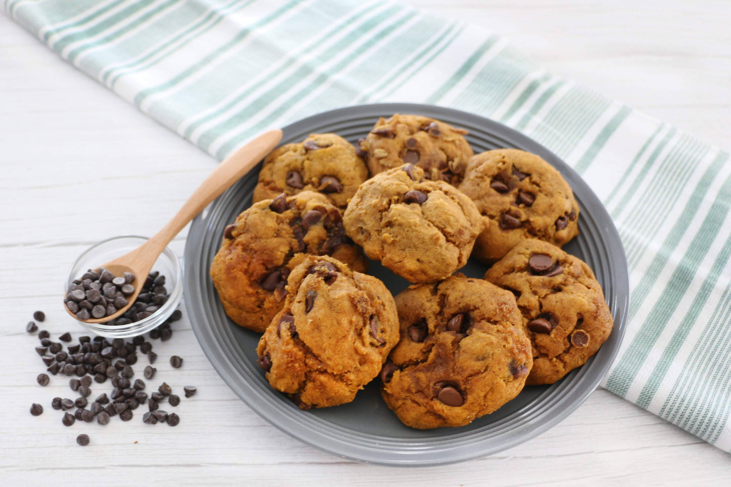 Plate of pumpkin chocolate chip cookies with a wooden spoon and scattered chocolate chips on a striped cloth.