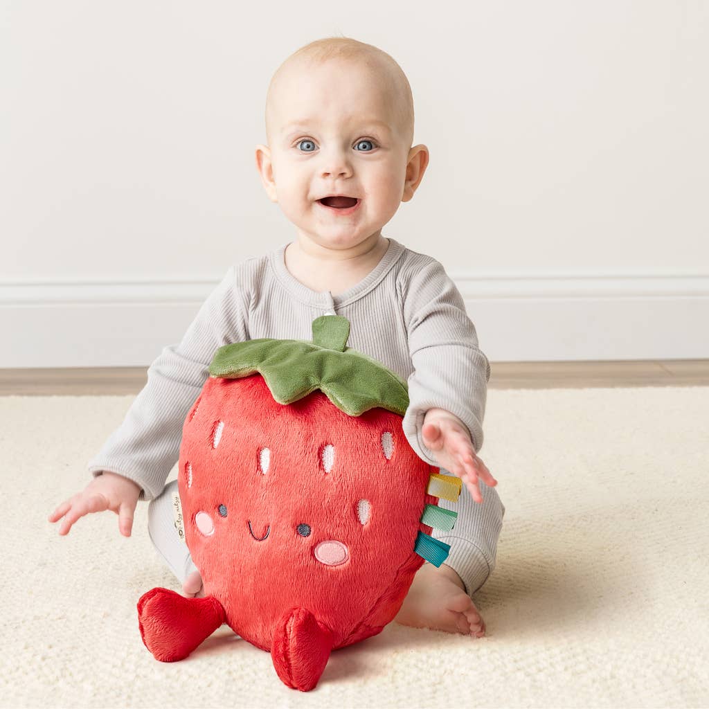 Baby holding a strawberry-shaped plush toy on a light-colored floor.