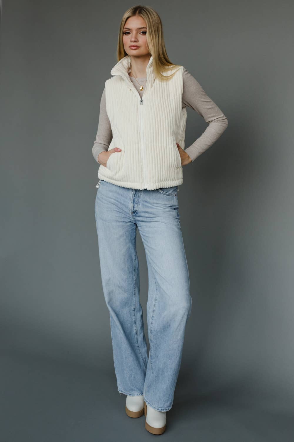 Woman wearing a cream vest, light gray long-sleeve shirt, blue jeans, and white shoes against a gray background