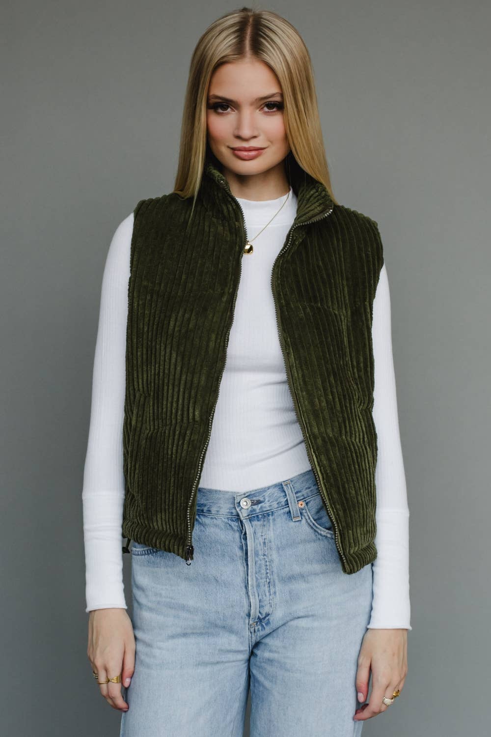 Woman wearing a green corduroy vest over a white shirt and blue jeans on a gray background