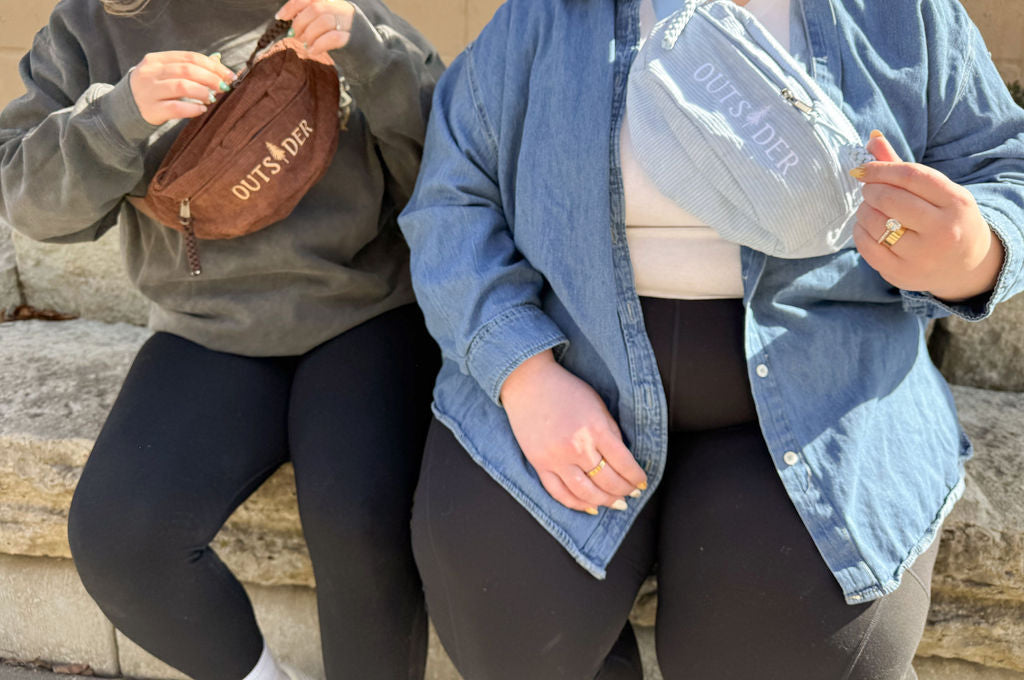Two women in black leggings with denim shirt or green sweatshirt wearing brown and blue corduroy fanny pack belt bags with the word Outsider embroidered on it in front of a stone wall