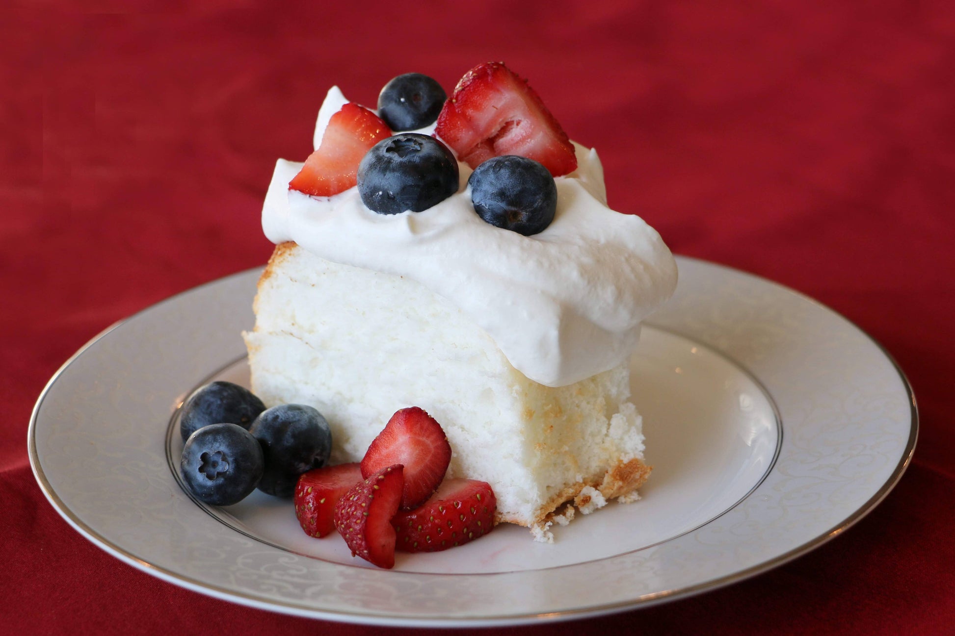 Slice of angel food cake with whipped cream and berries on a plate against a red background