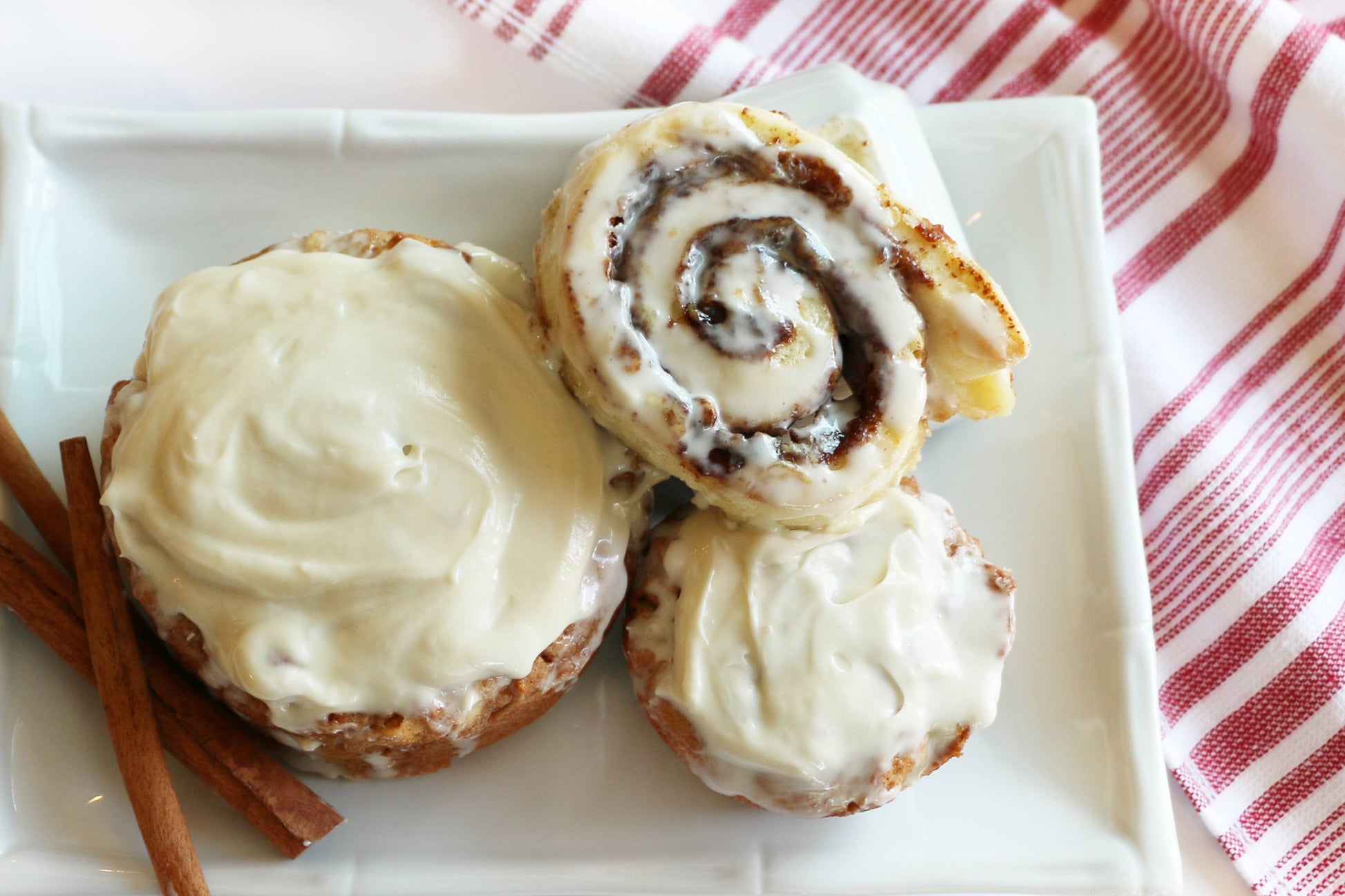 Cinnamon rolls with cream cheese frosting on a white plate with cinnamon sticks and a red and white striped towel.