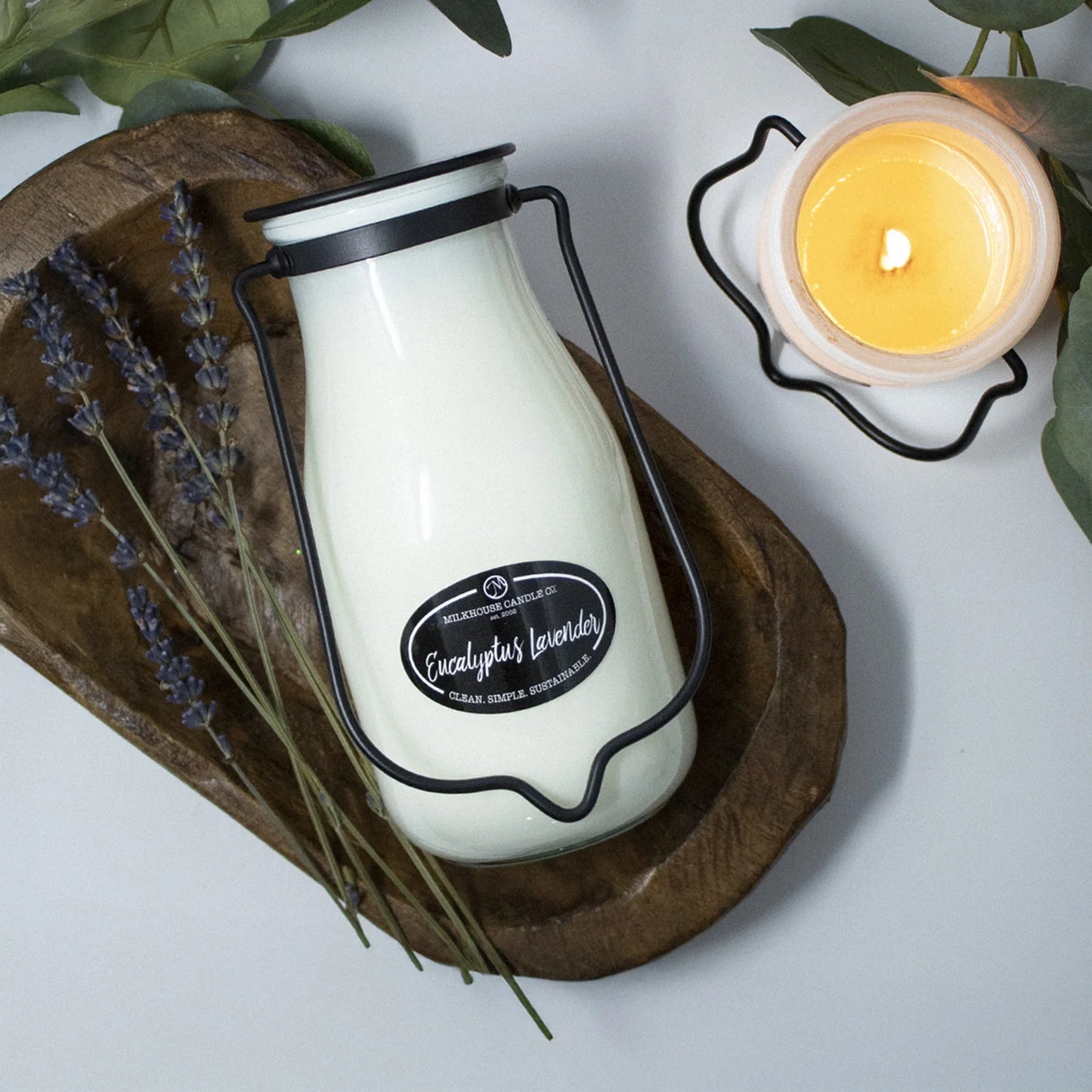 White candle in a glass jar with black lid on a wooden board, surrounded by lavender and a lit candle.