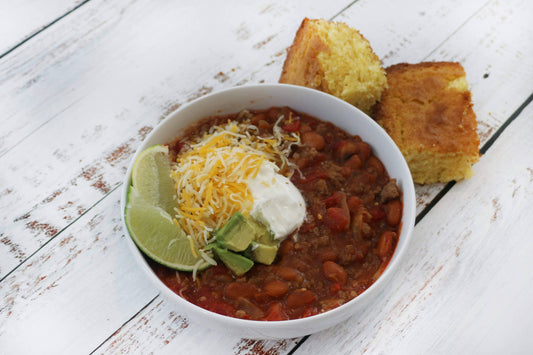 Bowl of chili with cornbread, sour cream, cheese, and a lime wedge on a wooden surface.