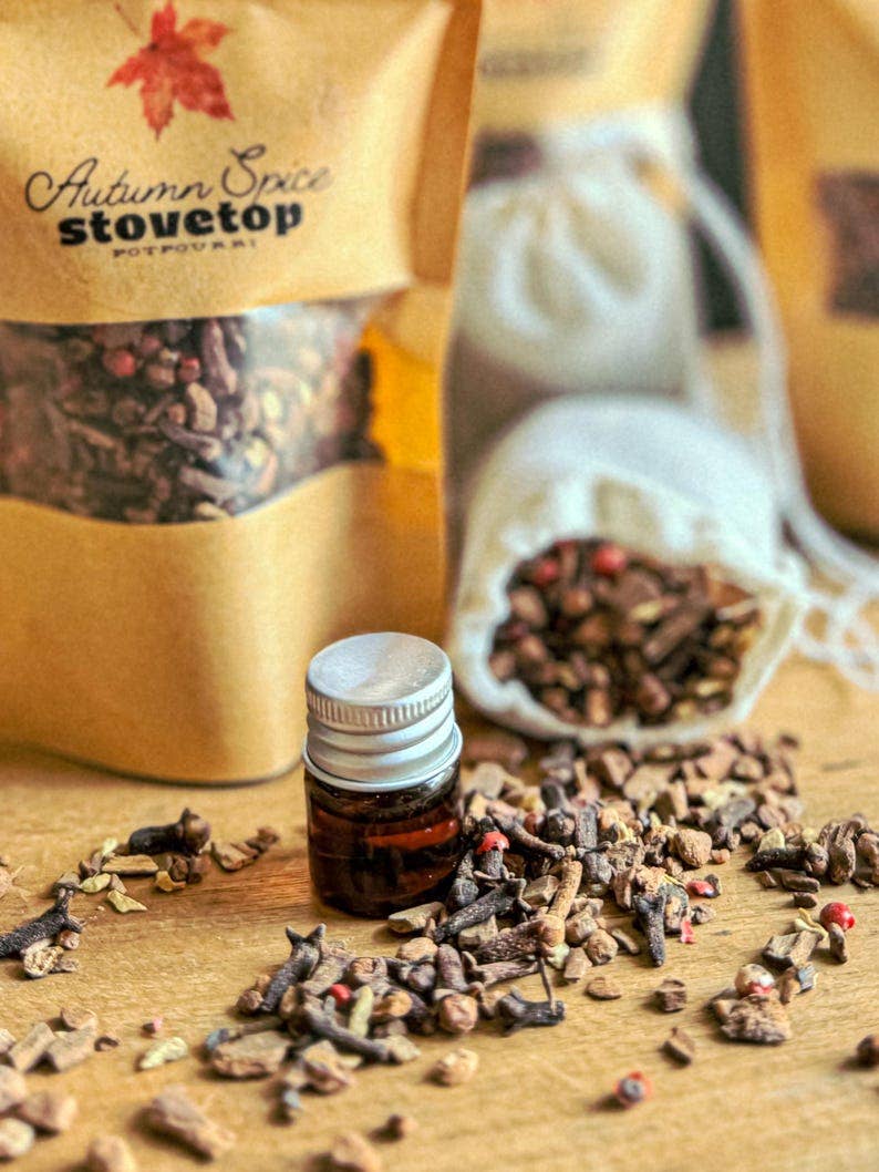 Autumn Spice Stovetop potpourri in a kraft paper bag with a clear window, shown alongside a small amber glass bottle with a silver cap and loose potpourri scattered on a wooden surface.