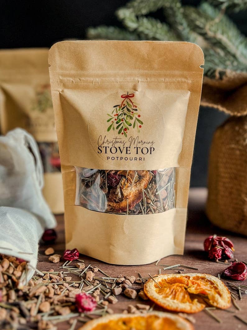 A kraft paper stand-up pouch labeled 'Christmas Wreath Stove Top Potpourri' with a clear window showing dried botanicals inside, styled on a rustic wooden surface with scattered dried flowers, tea bags, and greenery in the background.