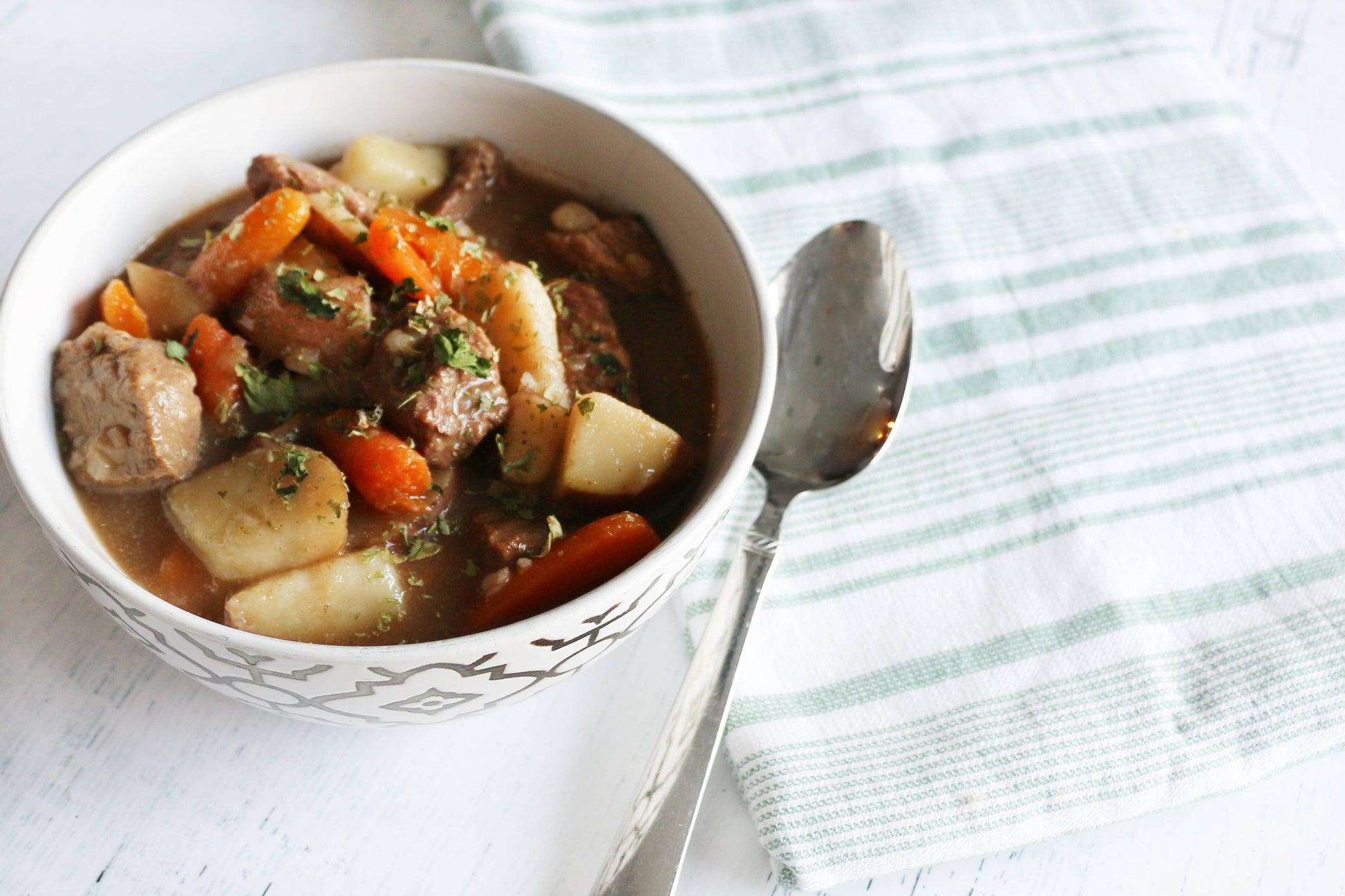 Bowl of beef stew with carrots and potatoes on a white surface with a striped cloth.