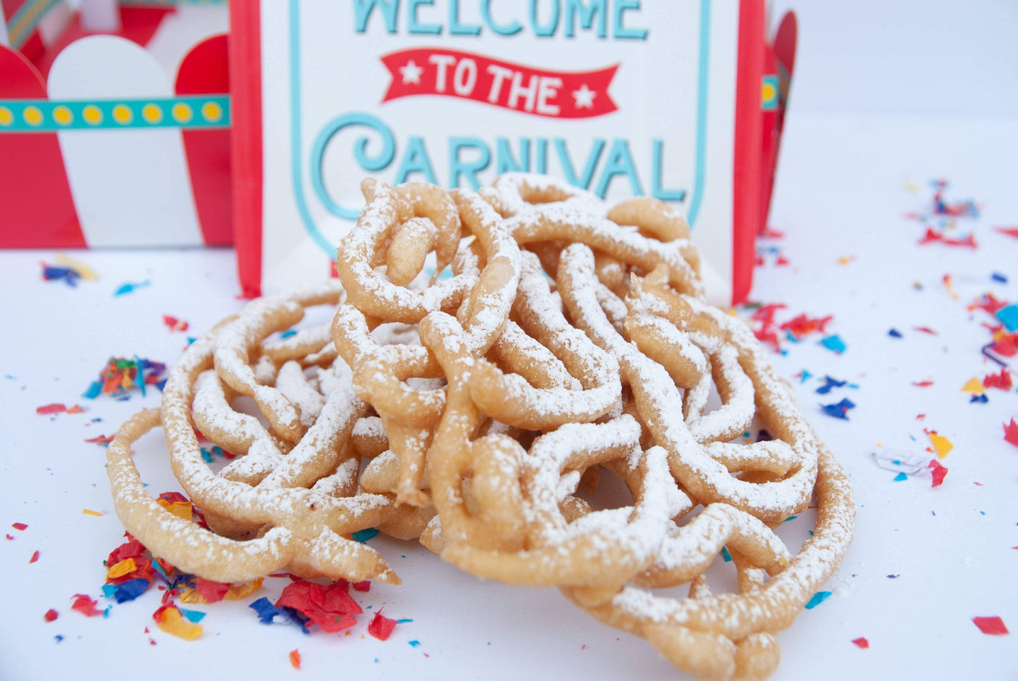 Funnel cake twists with powdered sugar in front of a 'Welcome to the Carnival' sign.