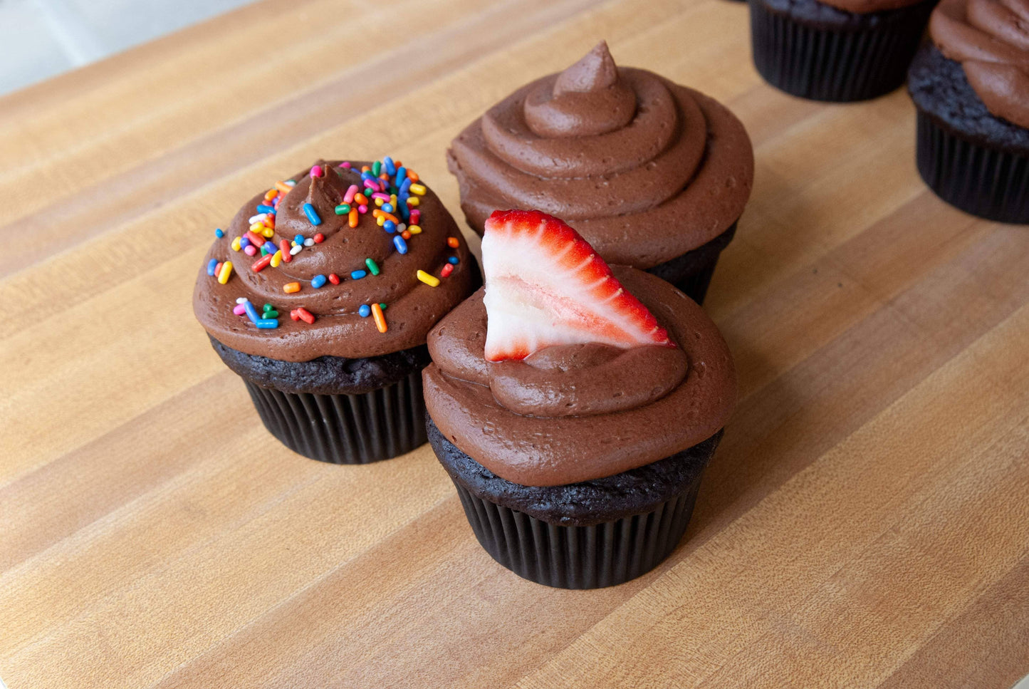 Chocolate cupcakes with chocolate frosting, sprinkles, and a strawberry on a wooden surface.
