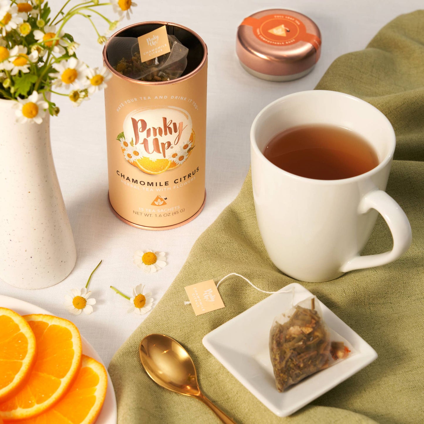 Pinky Up chamomile citrus tea canister with a cup of tea, orange slices, and a spoon on a green cloth.