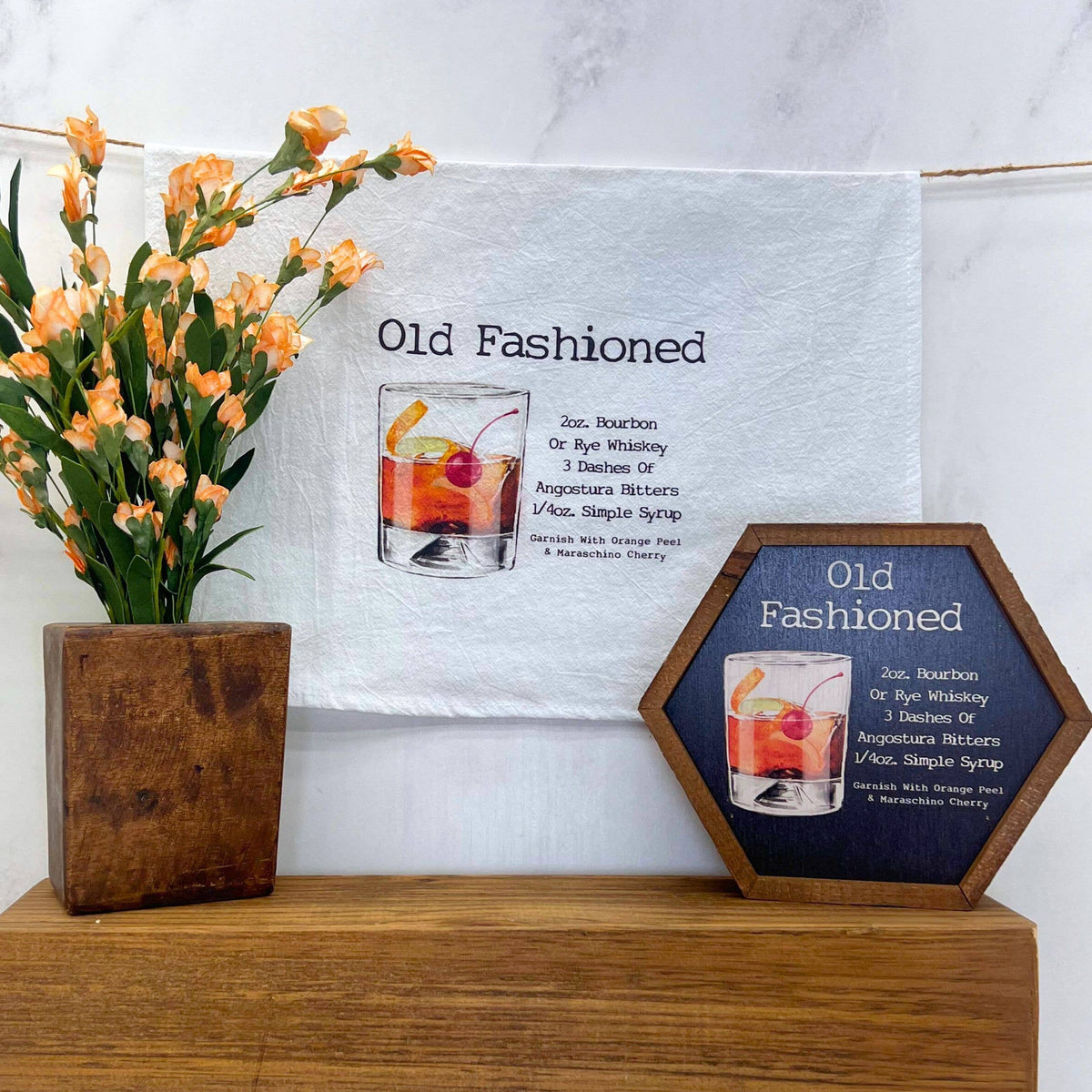 Decorative signs with 'Old Fashioned' cocktail tea towel and illustration on a wooden surface with flowers.