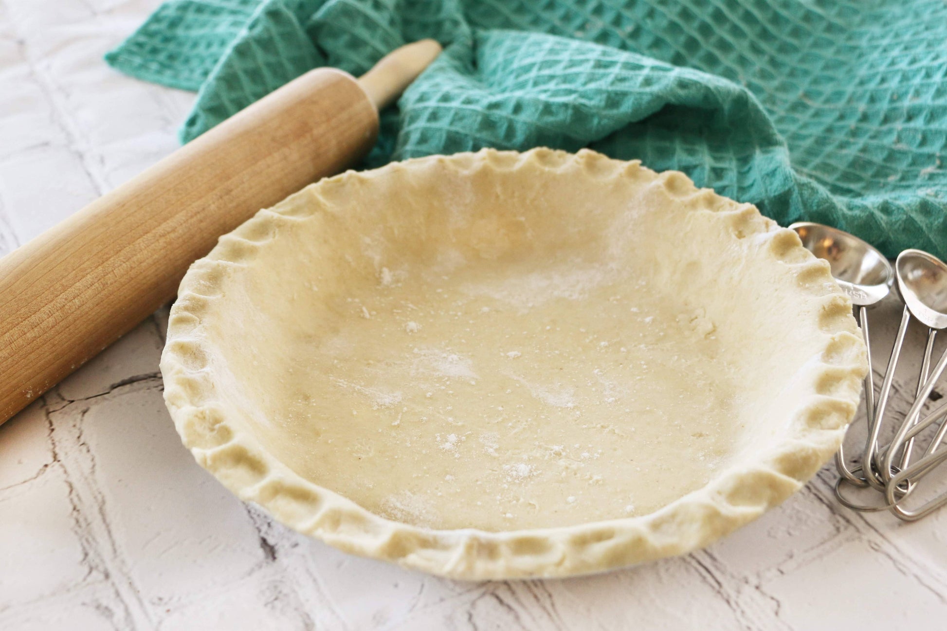 Unbaked pie crust on a white surface with a rolling pin and green towel in the background.