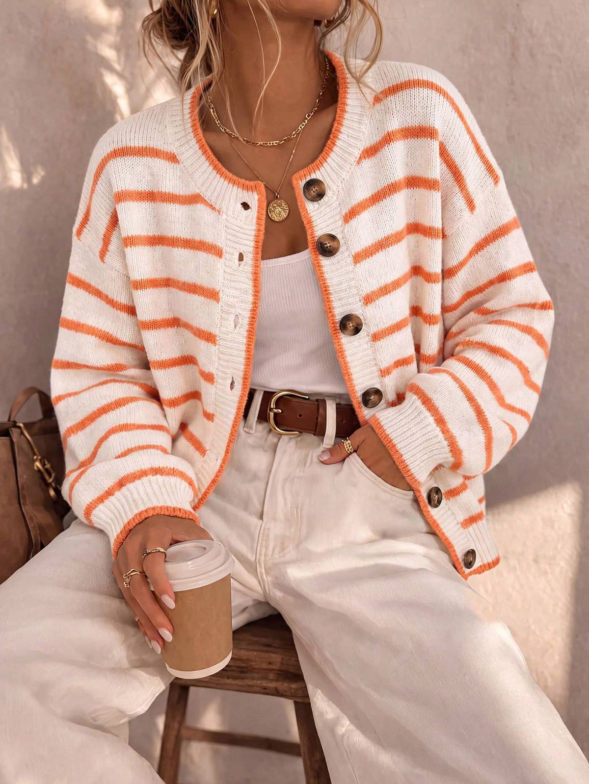 A woman seated on a stool wearing a cream knit cardigan with bright orange horizontal stripes, brown buttons, and a white tank top with white pants. She is holding a takeaway coffee cup in a warm, sunlit indoor setting.