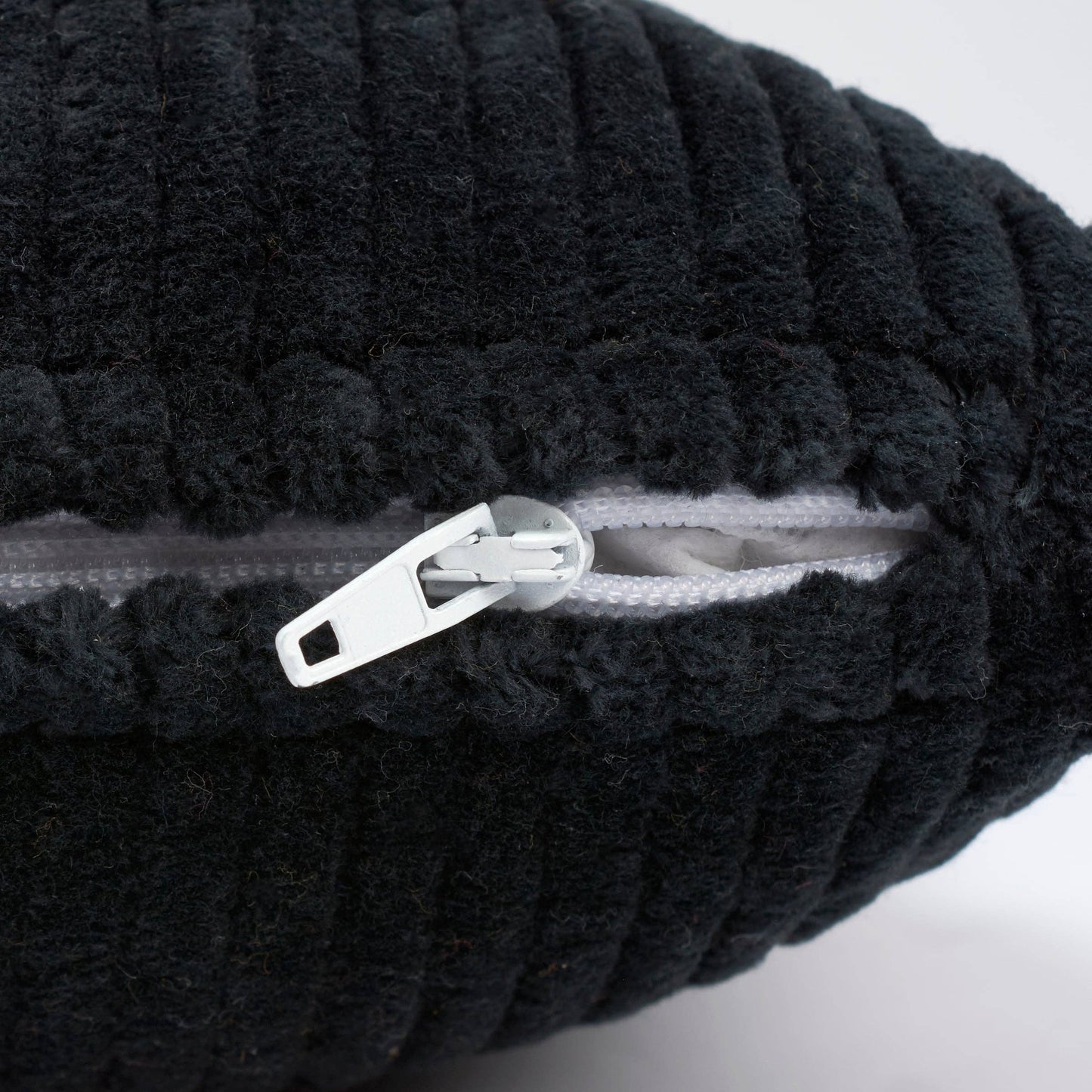 Close-up of a black ribbed plush or bouclé fabric item with a white zipper and white zipper pull, shown against a light neutral background.