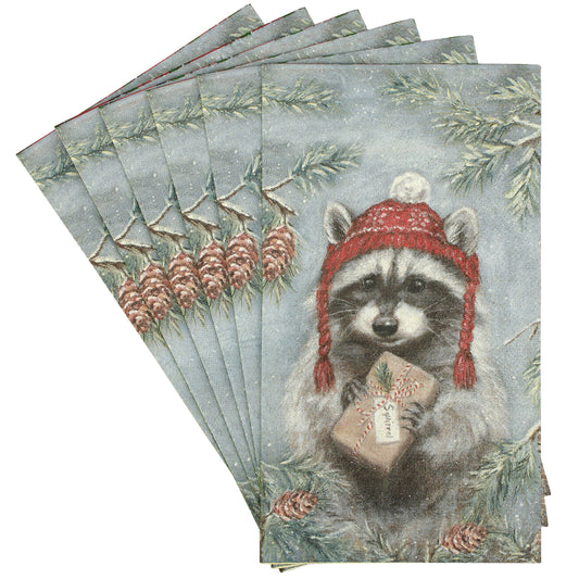 A fan of rectangular paper napkins featuring a vintage-style illustration of a raccoon wearing a red knit hat and holding a wrapped gift, surrounded by pine branches and pinecones on a pale blue-gray background.