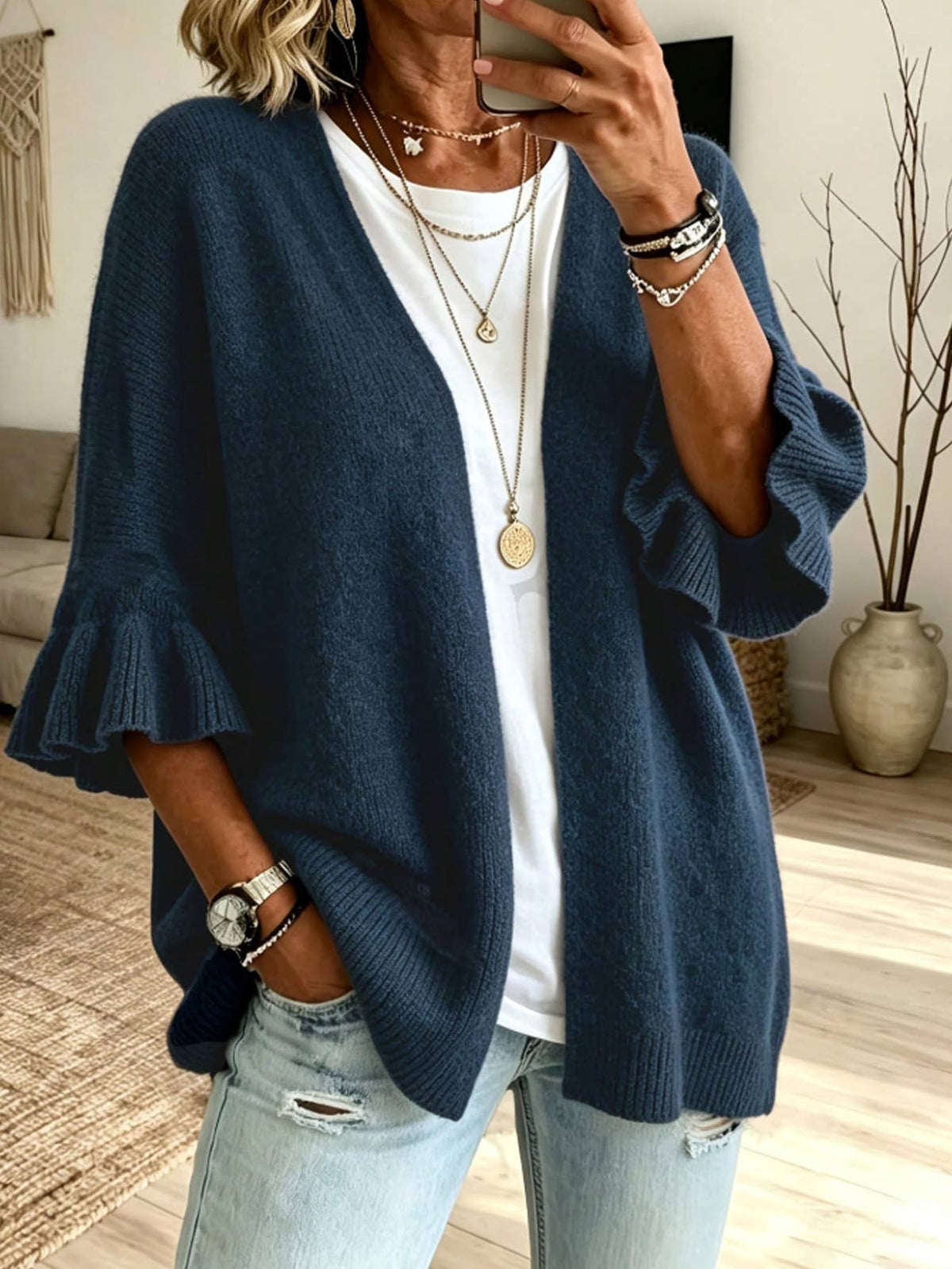 A woman takes a mirror selfie wearing a navy blue open-front cardigan with ruffled bell sleeves over a white t-shirt and light-wash distressed jeans. She is accessorized with layered gold necklaces, bracelets, and a watch.