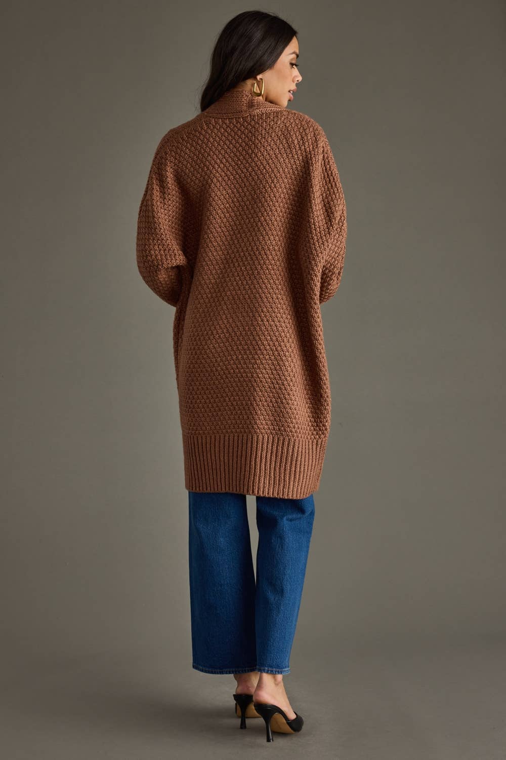 Woman wearing a brown knitted cardigan and blue jeans on a gray background