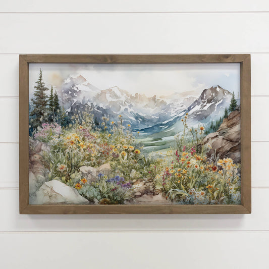 Framed artwork of a mountain landscape with flowers and trees on a white wall.
