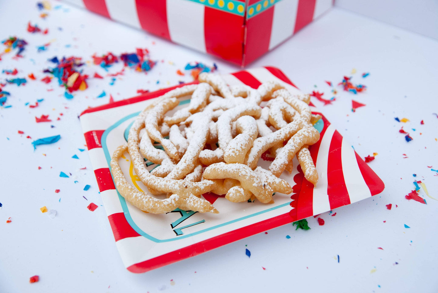 Funnel cake on a red and white striped plate with colorful sprinkles on a white background