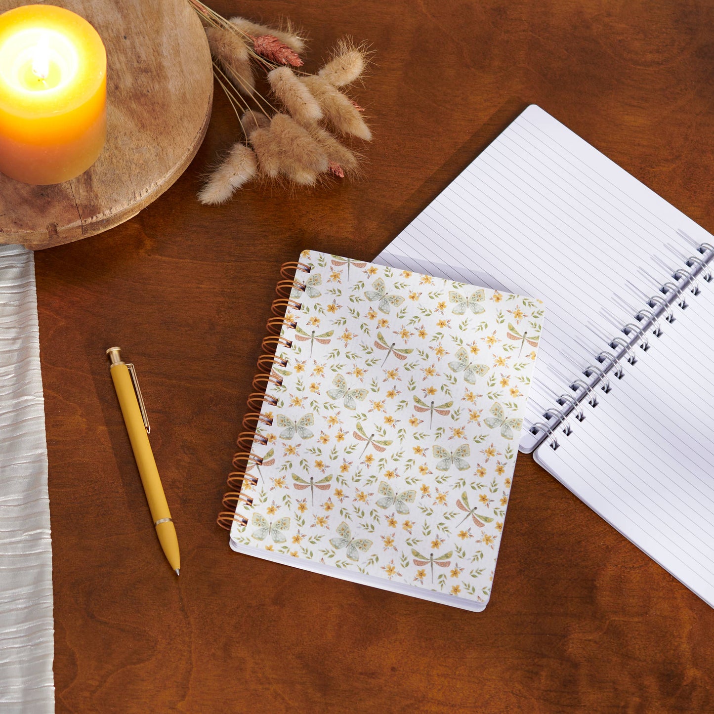 Floral notebook, pen, and open notebook on a wooden surface with a candle and dried plants.