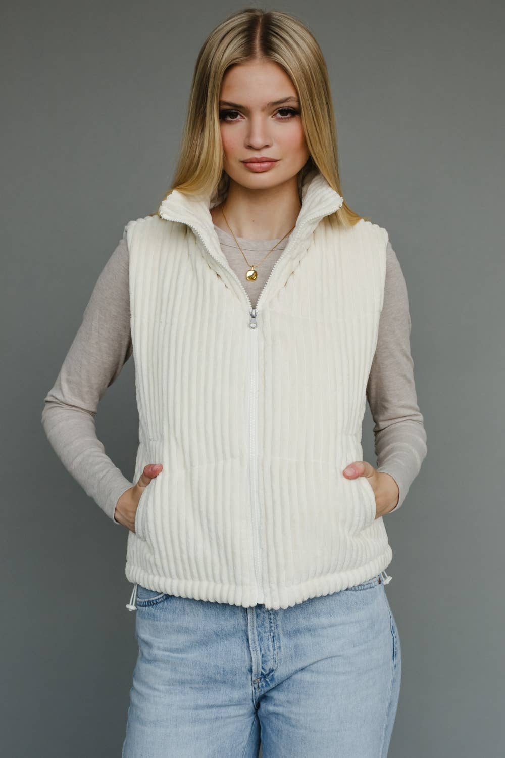 Woman wearing a cream ribbed vest over a gray long-sleeve shirt and blue jeans on a gray background